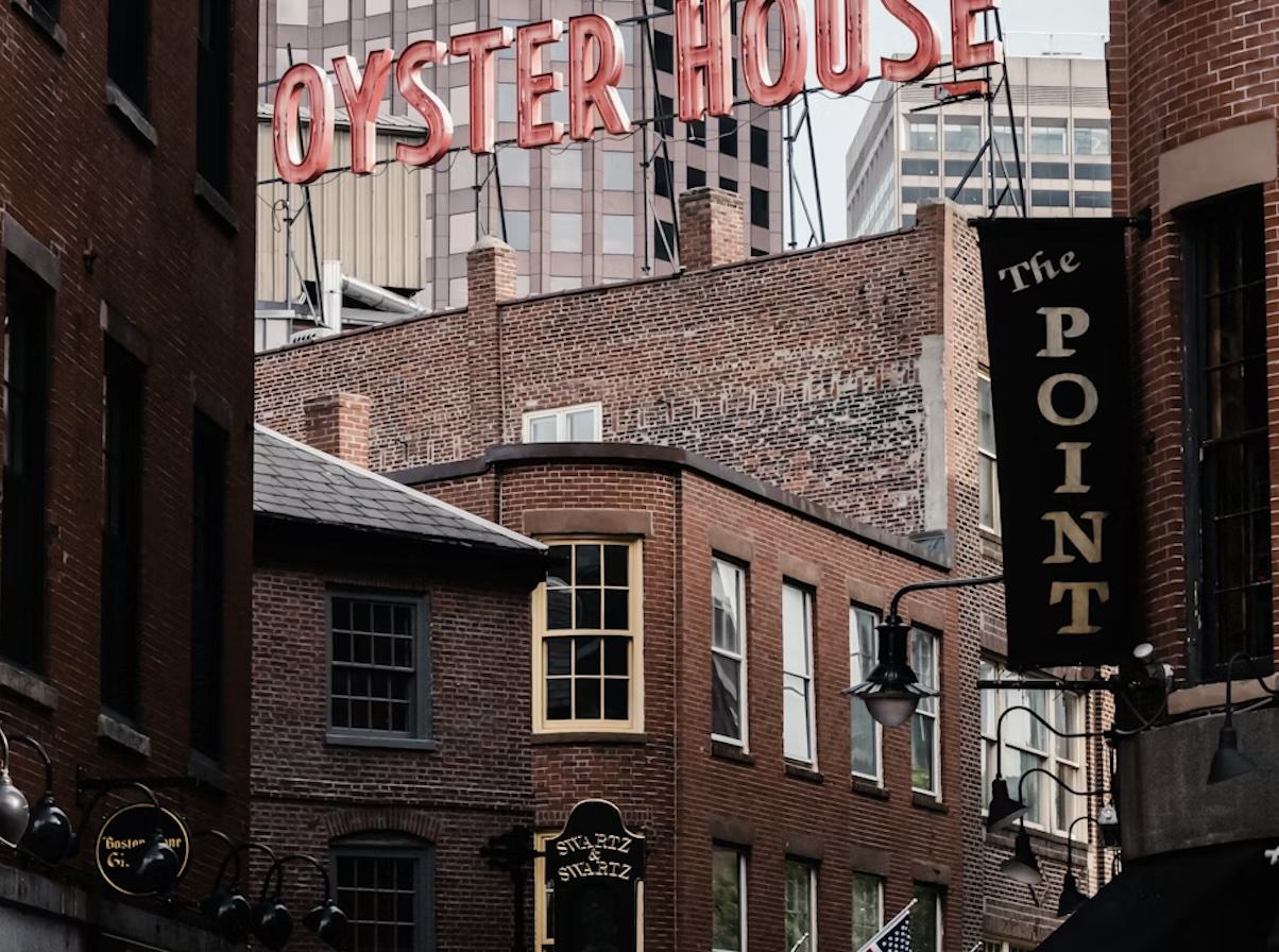 A close-up, urban shot of historic brick buildings in Boston's Blackstone Block. A large, red neon "OYSTER HOUSE" sign sits atop a building in the background, while a vertical black sign for "The POINT" hangs in the foreground. Narrow alleys and multi-pane windows emphasize the colonial-era architecture.