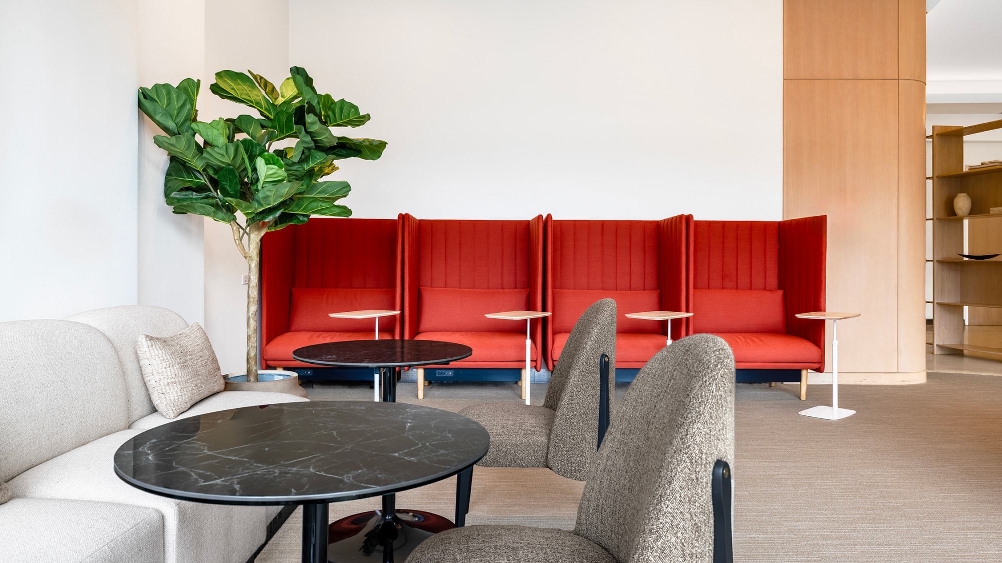 A modern lounge area with four high-backed red privacy booths featuring small, adjustable white laptop tables and built-in power outlets. In the foreground are black marble-topped cafe tables, a beige sofa, and a large potted fiddle-leaf fig plant.