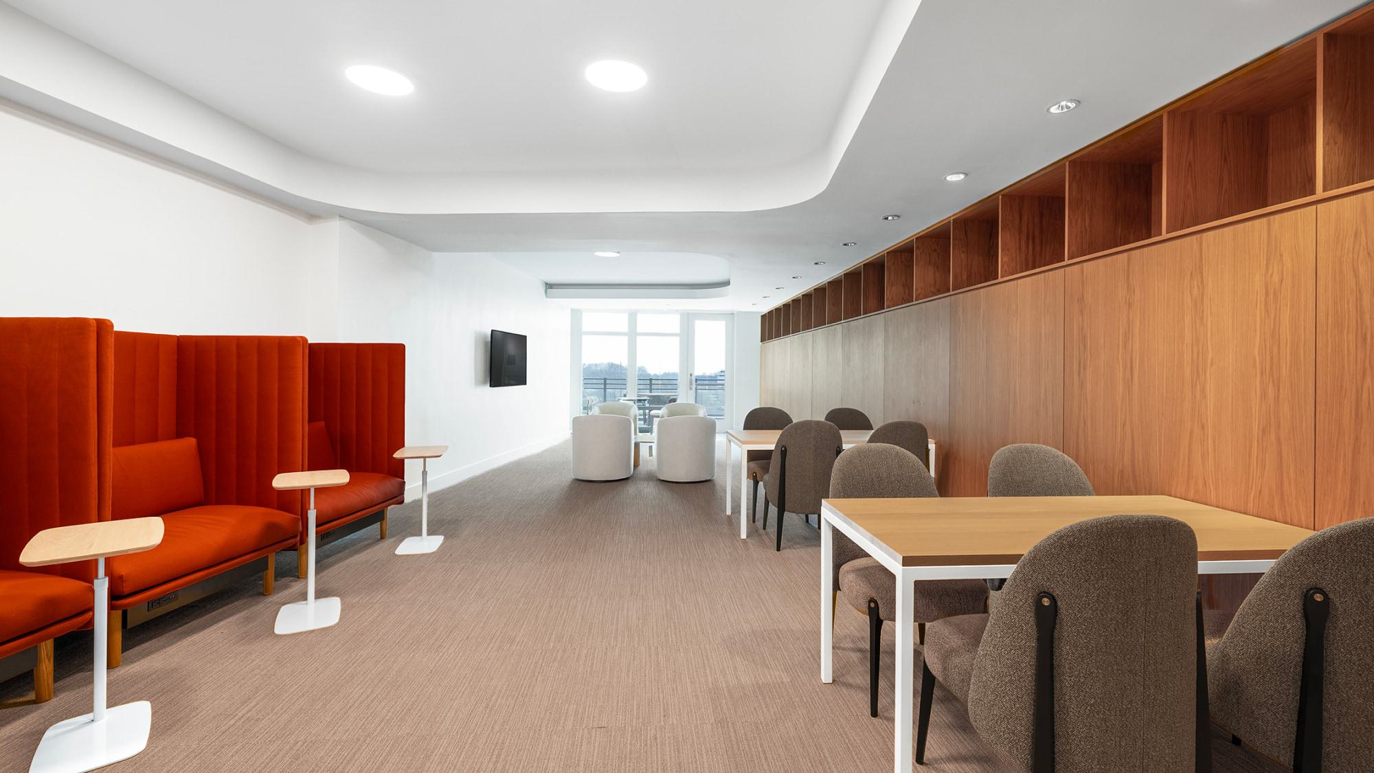 A bright, minimalist coworking space with several wooden work tables and grey upholstered chairs. On the left, two tall orange privacy booths with adjustable side tables sit against a white wall, and a long wood-paneled storage unit runs along the right wall.