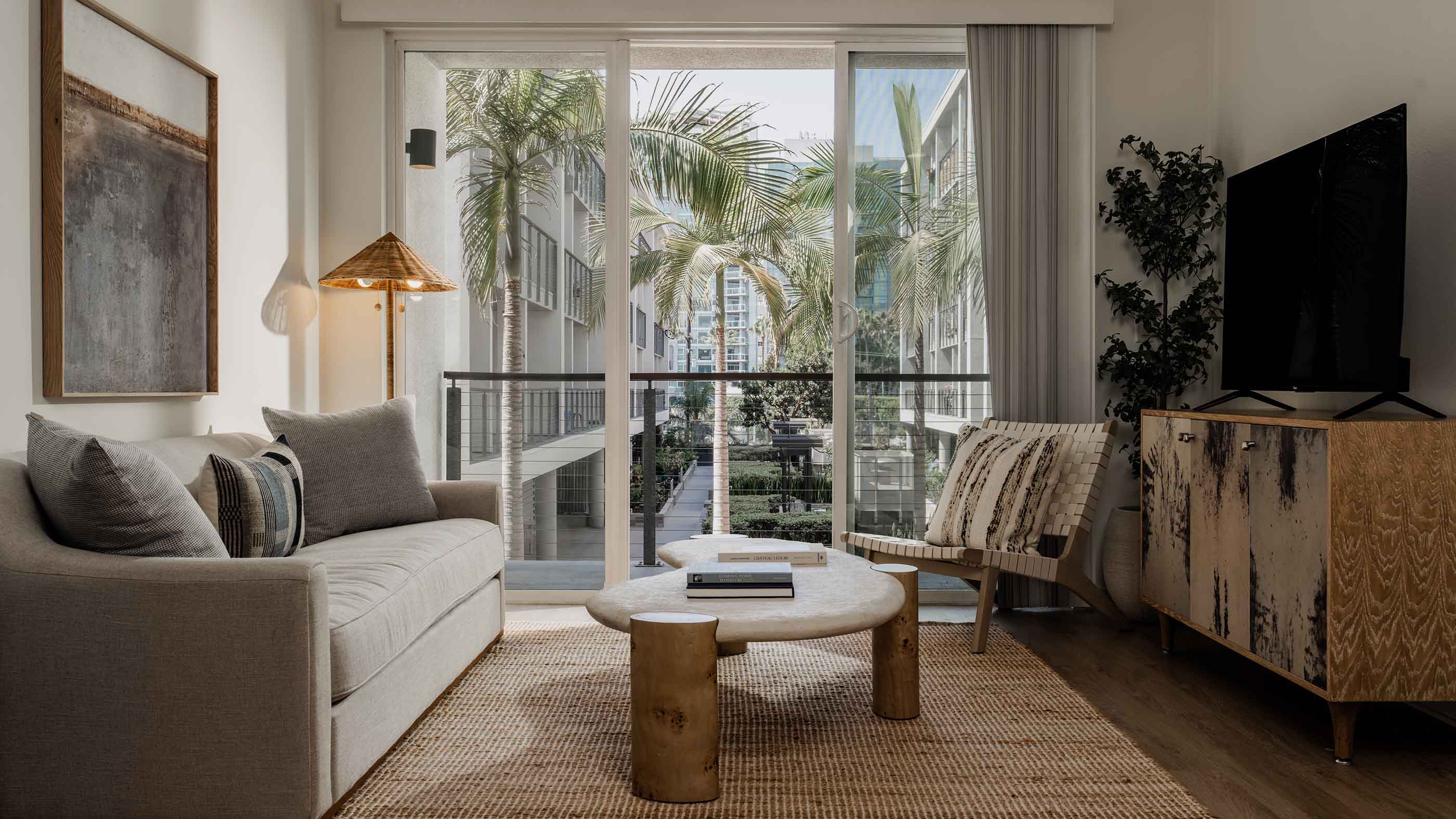 Sunlit townhome living area with low-profile beige seating, round marble coffee table, and a potted olive tree.