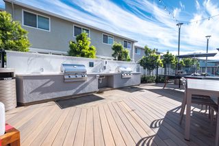 A modern outdoor communal kitchen on a wood-plank deck featuring built-in stainless steel grills, sleek stone-inspired countertops and backsplash, a prep sink, and a communal dining table.