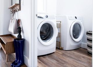 A bright laundry area featuring side-by-side white front-load washer and dryer units on wood-style flooring, adjacent to a modern entryway with a wooden bench and coat hooks.