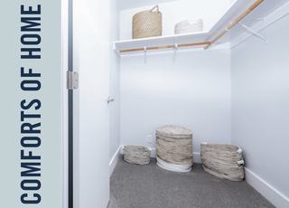 A spacious walk-in closet with plush grey carpeting, white walls, and built-in shelving with wooden hanging rods. The space is styled with various woven storage baskets on the floor and top shelf.
