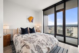 A modern high-rise bedroom at Atlas Oakland featuring floor-to-ceiling windows and a glass door leading to a private balcony with panoramic city views. The room is styled with a patterned duvet and contemporary lighting.