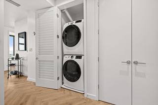 A modern high-rise apartment hallway featuring warm herringbone wood flooring and a closet with a louvered door opened to reveal a white, stacked front-load washer and dryer.