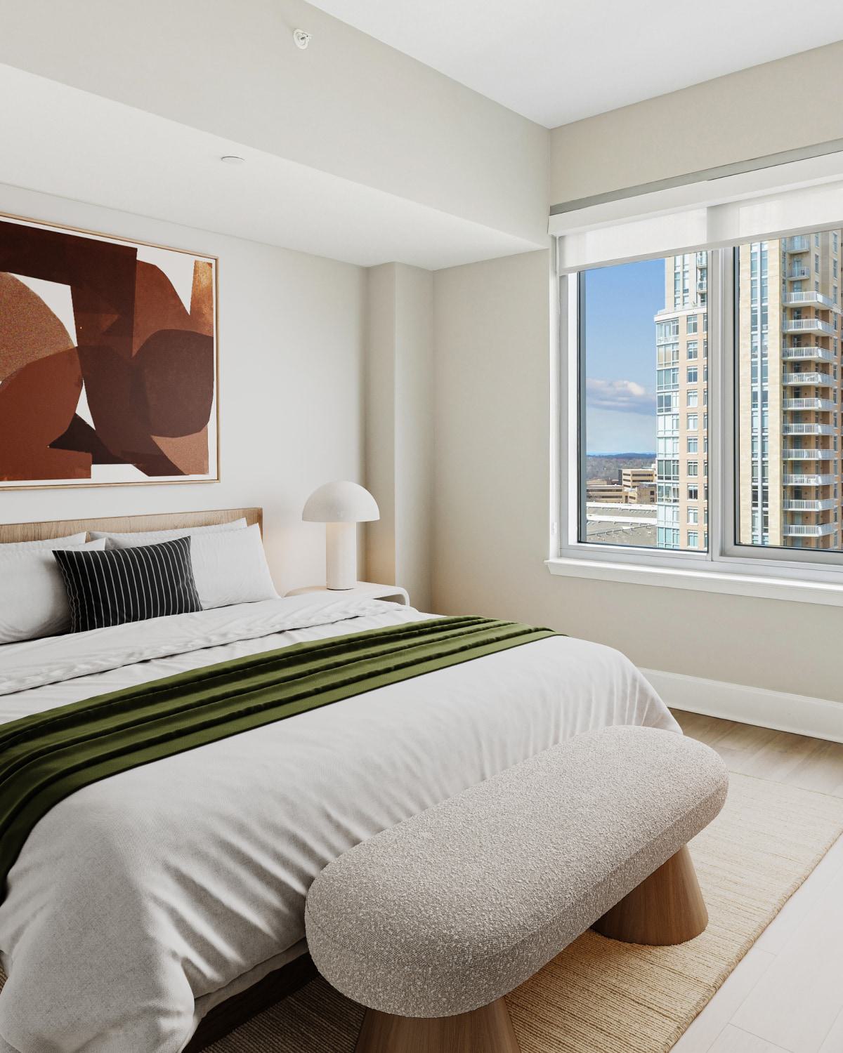 Modern apartment bedroom featuring a large window with Reston Town Center views, a neatly made bed with white linens and an olive-green throw blanket, abstract wall art, and mushroom-shaped bedside lamps.