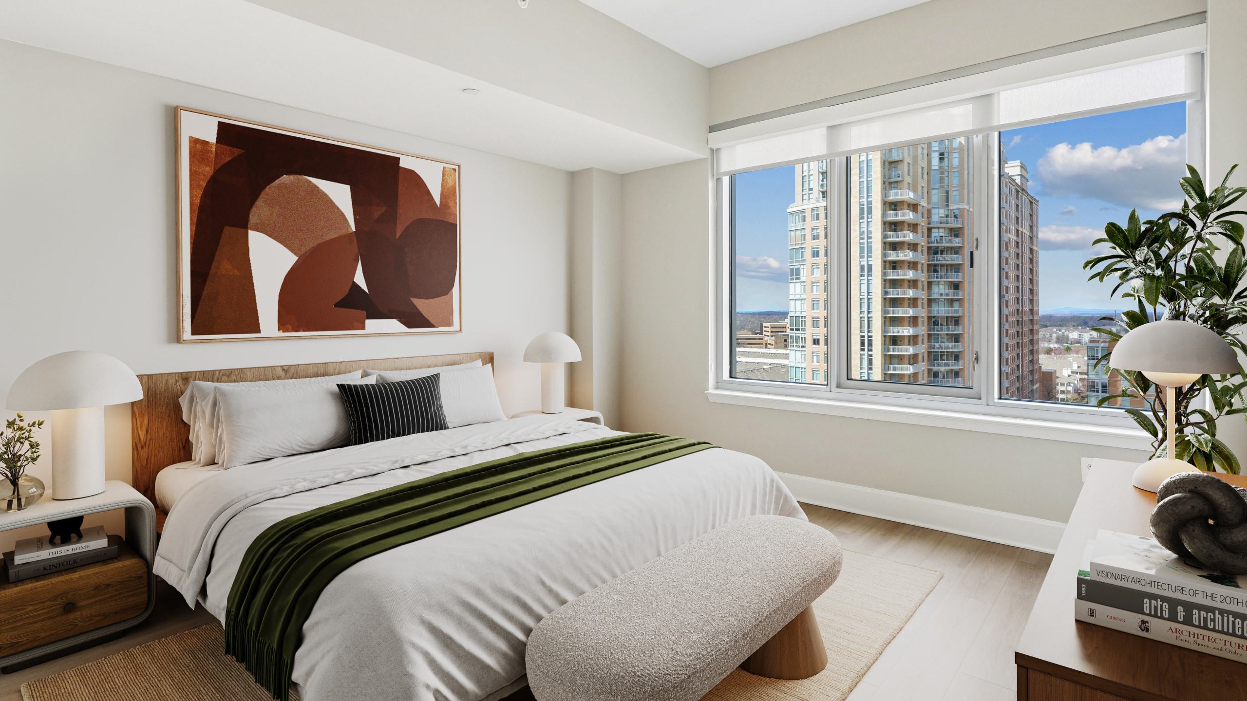 Modern apartment bedroom featuring a large window with Reston Town Center views, a neatly made bed with white linens and an olive-green throw blanket, abstract wall art, and mushroom-shaped bedside lamps.