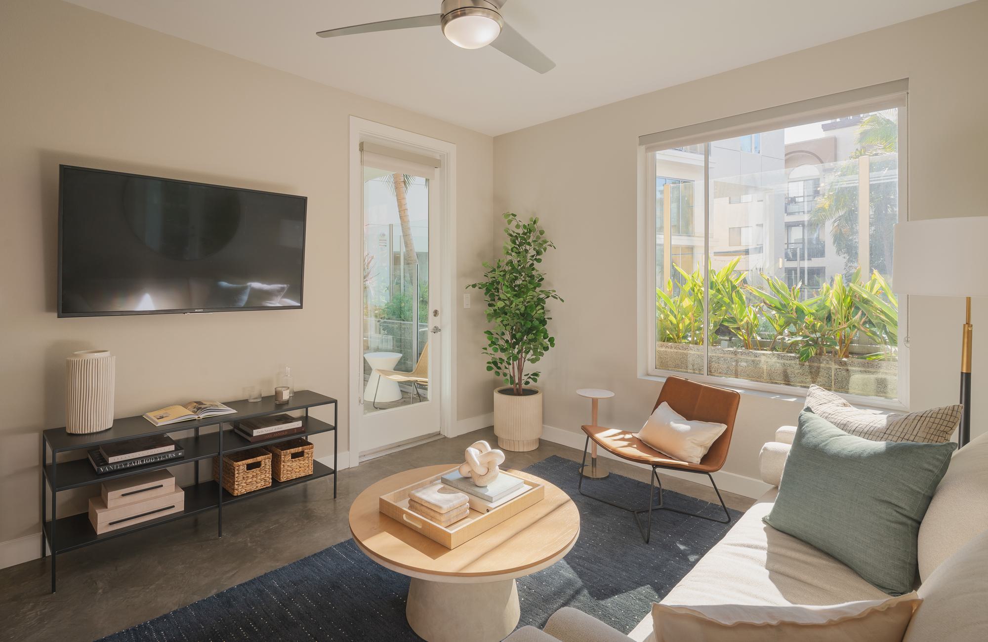 Bright apartment living room with concrete floors, a round wooden coffee table, and a large window overlooking lush tropical plants and a private balcony.