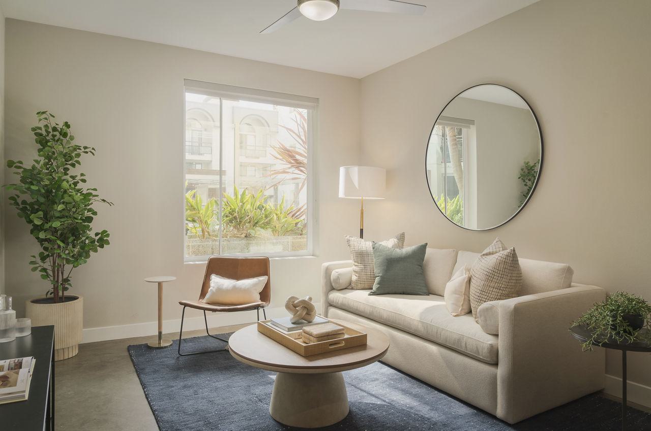 Modern living area featuring a large square window with neighborhood views, a cream sofa, and a large circular wall mirror reflecting natural light.
