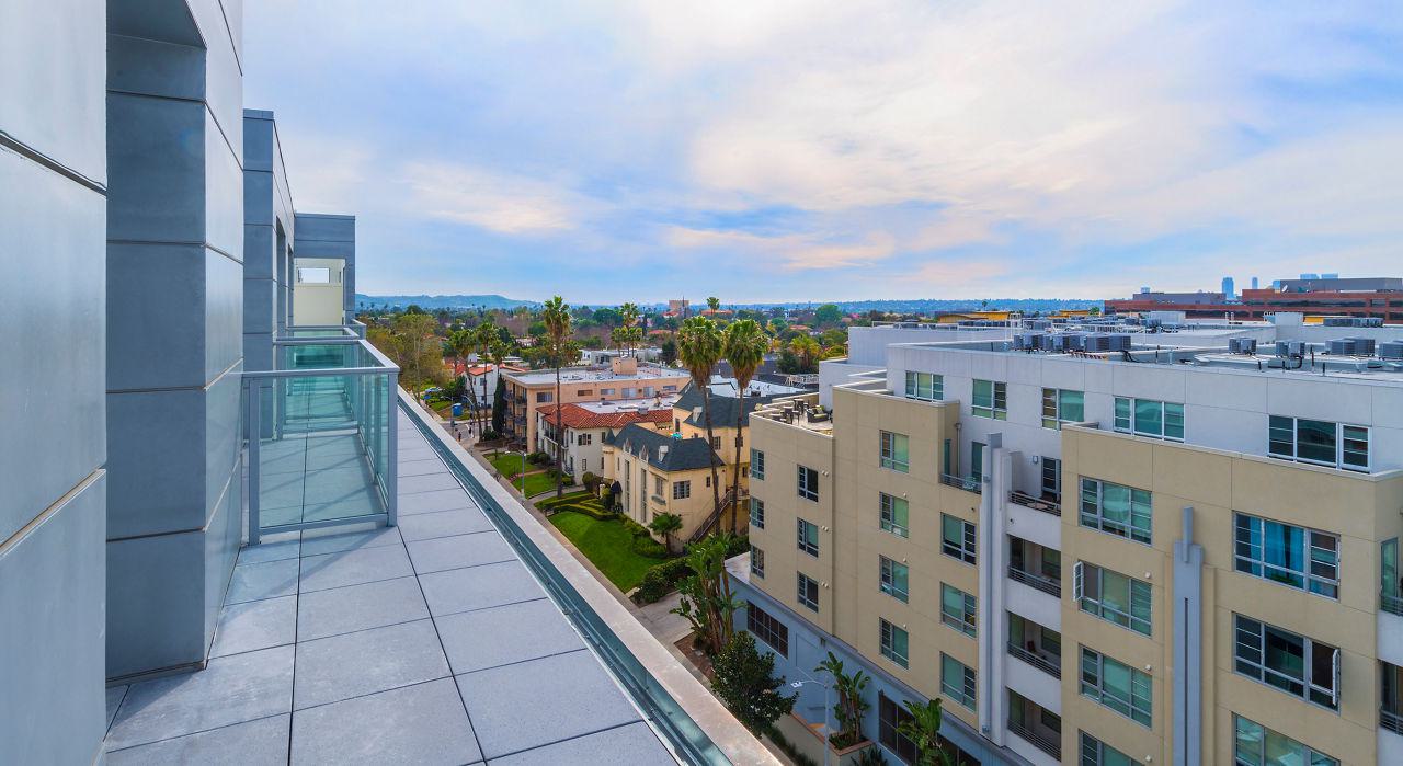An elevated view from a private upper-level balcony showing a long glass railing and a perspective of the Miracle Mile neighborhood's mid-rise architecture and palm trees under a bright sky.