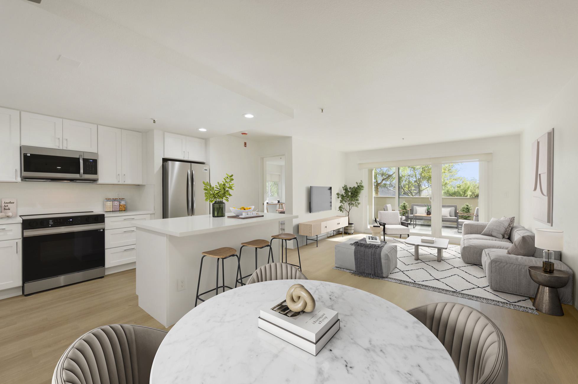 open-concept modern apartment featuring a bright white kitchen with stainless steel appliances, a breakfast bar with three stools, and a living area with a grey sectional sofa and a sliding glass door leading to an outdoor balcony area. In the foreground, a round marble dining table is partially visible.