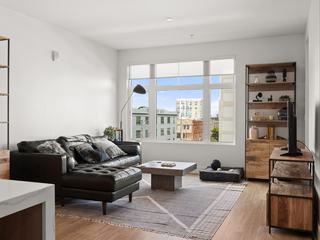 A bright, modern living room at Fourth Street East featuring a black leather sectional, wooden shelving, and a large oversized window showcasing a view of Jack London Square’s historic industrial buildings and city skyline.