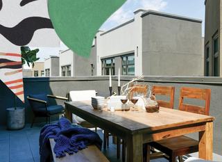 A spacious private rooftop balcony at Fourth Street East featuring a large wooden dining table set for a meal with wine and candles. The open-air space is surrounded by a low gray wall, offering a view of the neighboring building architecture under a bright sky.