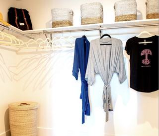 A detailed look inside a well-organized walk-in closet at Fourth Street East, featuring a wrap-around white wire shelf and hanging rods, a woven hamper on the wood floor, and curated items including a blue robe, a grey robe, and an "Oaklandish" logo t-shirt.