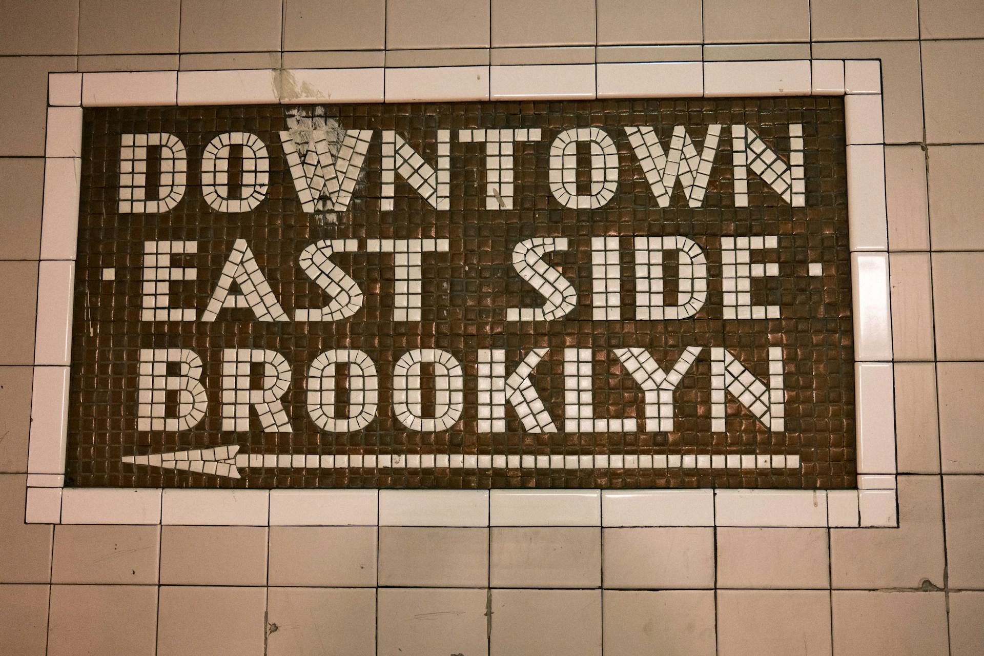 A vintage mosaic tile sign on a New York City subway station wall with a white border and brown background. The white tiled text reads 'DOWNTOWN EAST SIDE BROOKLYN' in a blocky, geometric font, with a white arrow pointing to the left.