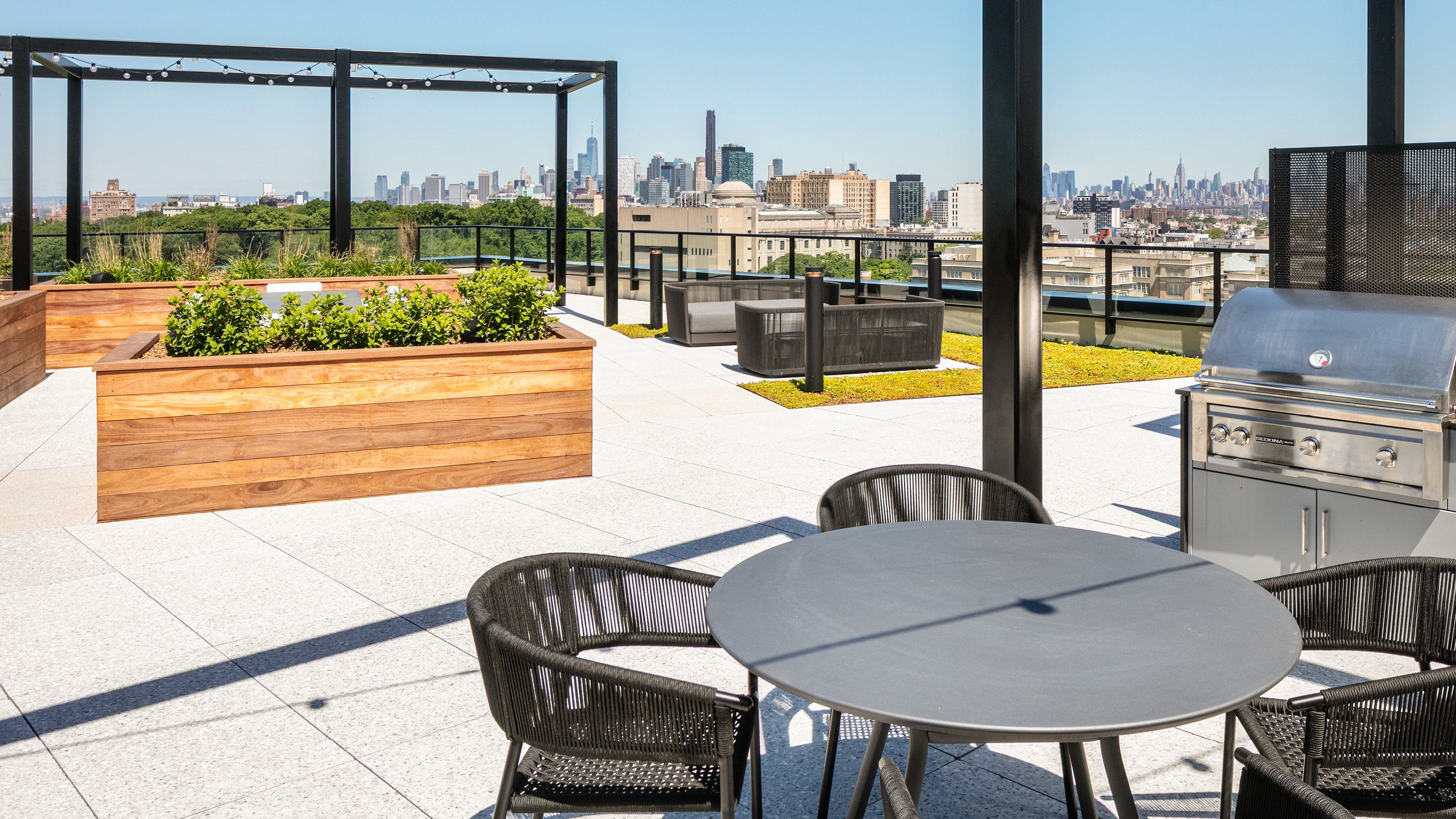 A spacious rooftop terrace offering panoramic views of the Manhattan skyline and the lush canopy of Prospect Park. The terrace is equipped with a stainless steel outdoor grill, a round dining table with black woven chairs, and built-in wooden planters filled with greenery. Grassy lawn patches and modern lounge seating are spread across the light gray stone pavers under a black architectural pergola.