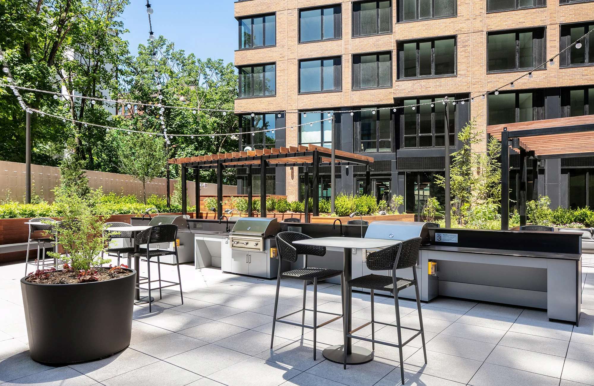 A sunny, modern outdoor amenity courtyard featuring a row of professional-grade stainless steel barbecue grills with gray stone prep counters. The space includes black woven high-top bistro tables and chairs, string lights draped overhead, and a large black planter. In the background, wooden pergolas and lush green trees border the building’s brick and black-windowed exterior.