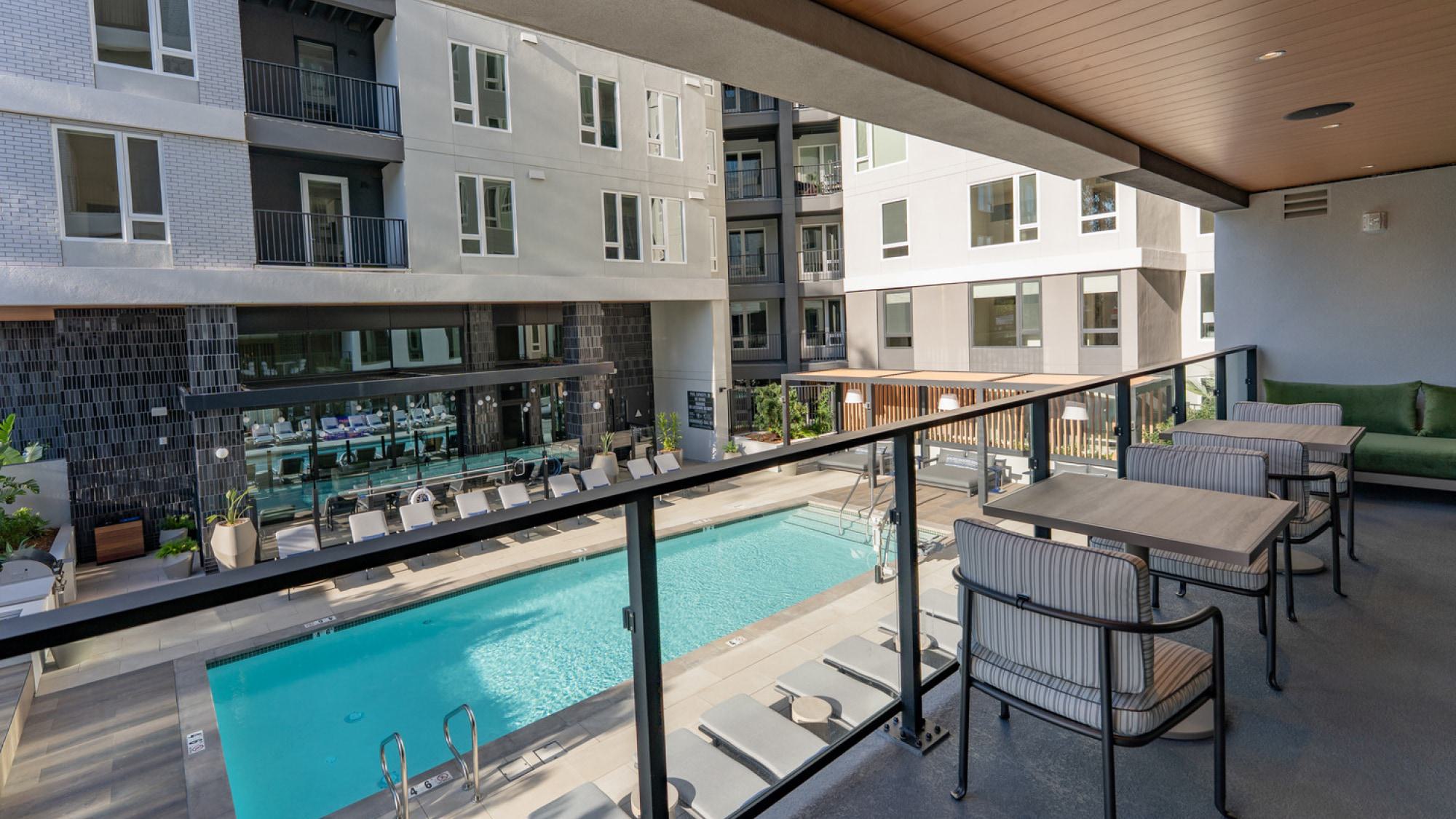 A dusk rendering of Revery, a multi-story modern apartment building in Burlingame. The exterior features a mix of dark grey panels, light brick, and wood-textured accents with numerous large windows and private glass-railed balconies. Manicured trees and a glow from the lobby windows line the street-level view.