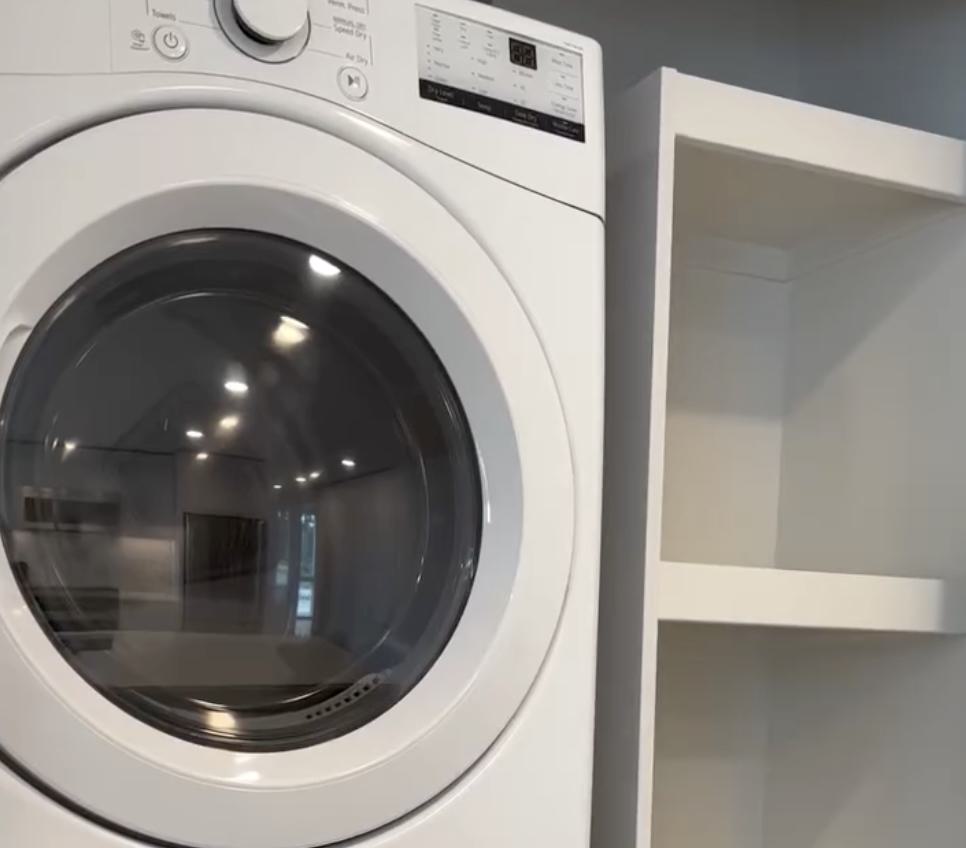 A close-up of a modern, white front-loading dryer featuring a digital display and circular glass door. The dryer is installed next to built-in white laundry room shelving, with the glass door reflecting the bright, contemporary interior of an apartment.