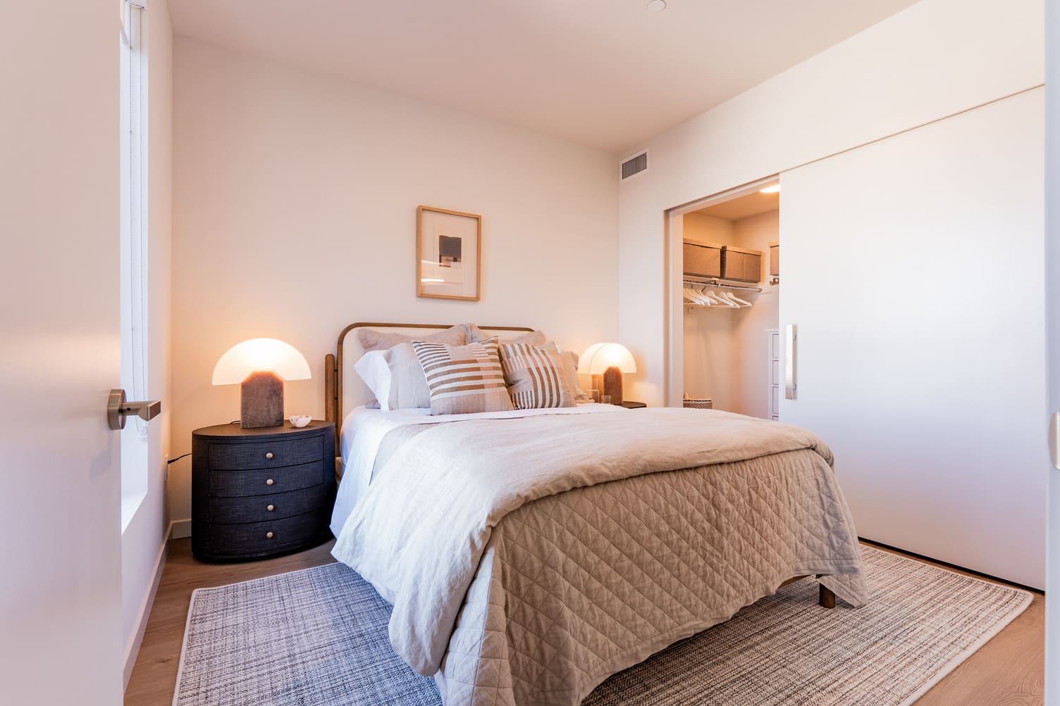 A bright, modern bedroom featuring a neutral-toned bed, warm bedside lamps, and a large sliding white door that opens to a walk-in closet. The closet interior shows an organized storage system with hanging rods and overhead shelving bins.