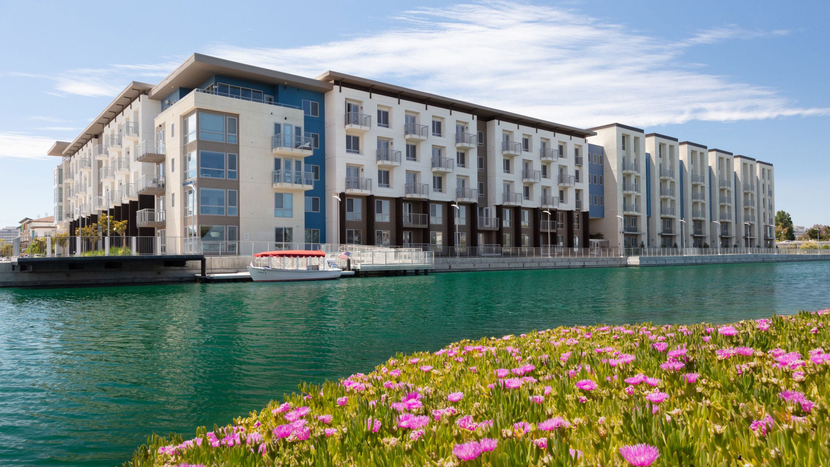 A wide-angle exterior view of The Triton luxury apartments in Foster City, situated along the tranquil lagoon. The building features a modern multi-story design with white and dark grey facades. The foreground shows a concrete waterfront promenade and a private boat dock reflecting in the calm blue water under a clear sky.