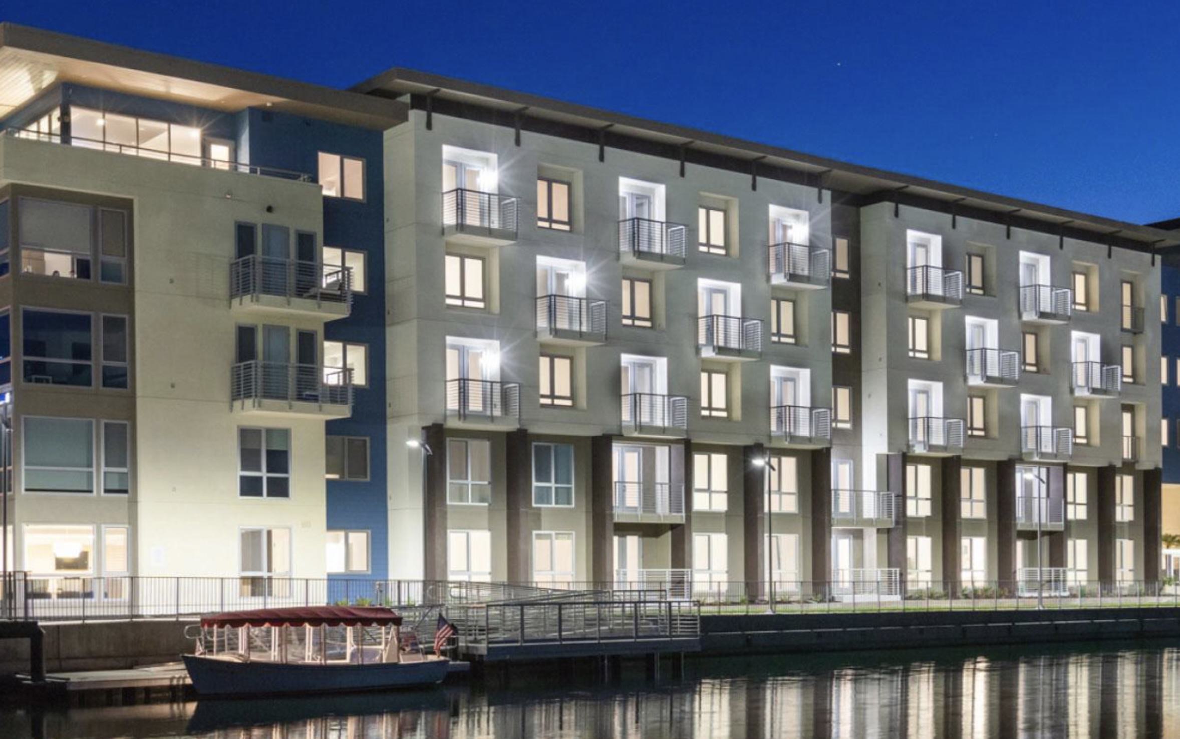 A twilight view of The Triton’s modern exterior. Warm light glows from apartment windows and private balconies, reflecting off the calm blue water of the lagoon. A red-canopied electric boat is moored at the private dock in the foreground.