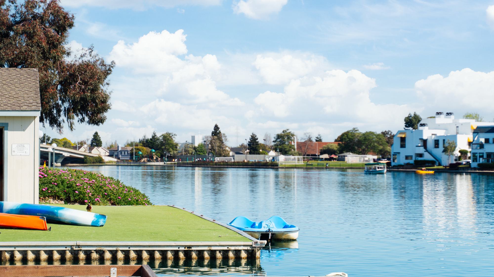 A bright, sunny day at the Foster City lagoon featuring a calm, blue waterway under a sky with fluffy white clouds. In the foreground, a green launching dock holds a bright orange kayak and a blue-and-white kayak, with a blue pedal le floating nearby. The background shows waterfront homes, lush green trees, and a low-profile bridge crossing the water.