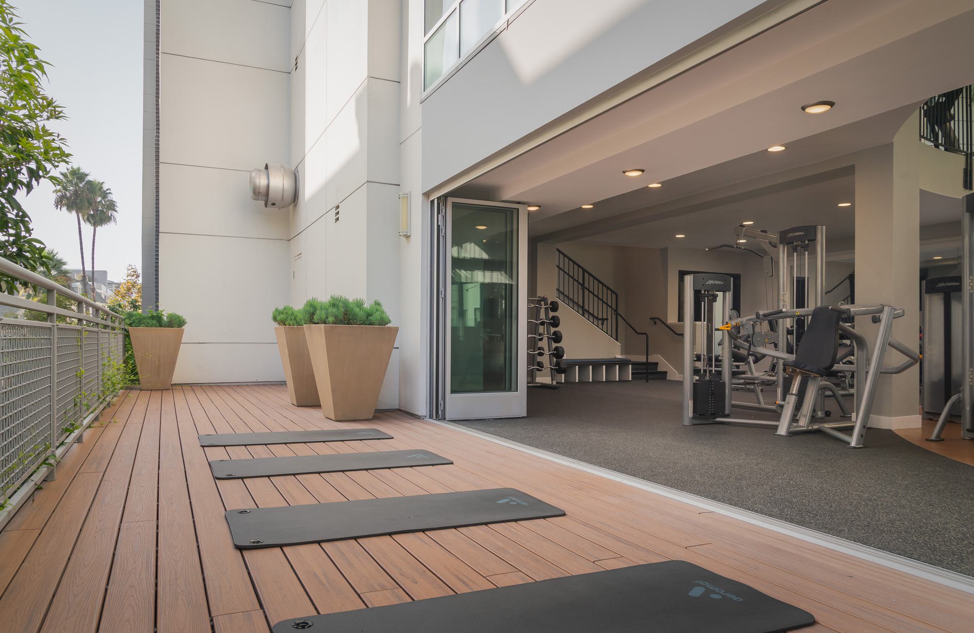An outdoor fitness and yoga deck with wood plank flooring and four black exercise mats laid out in a row. Large bi-fold glass doors open completely into an indoor gym area featuring strength-training equipment.