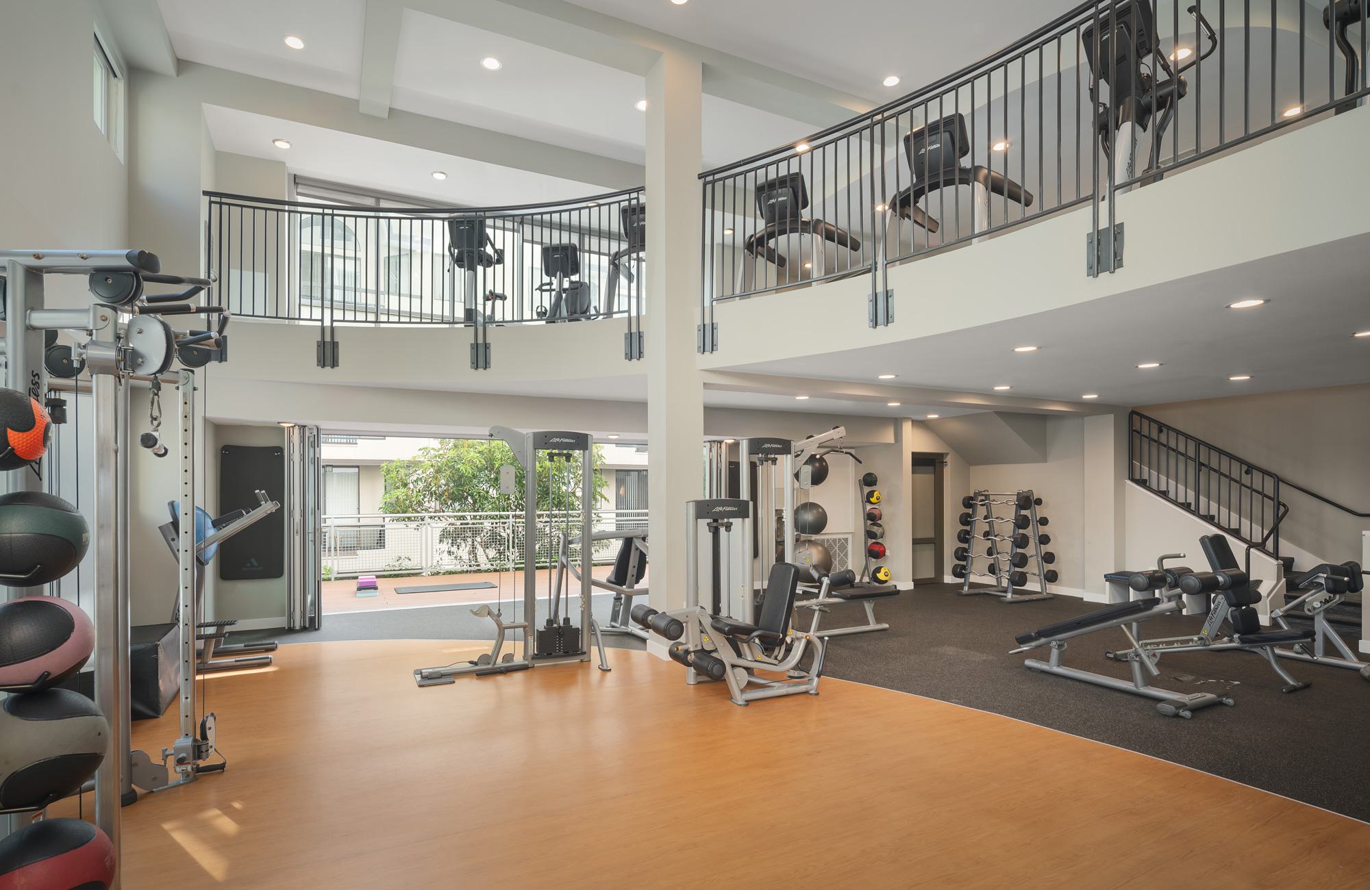 A spacious, multi-level indoor fitness center with a wood-look floor, a dedicated weight training area, and a second-story mezzanine balcony lined with cardio machines.