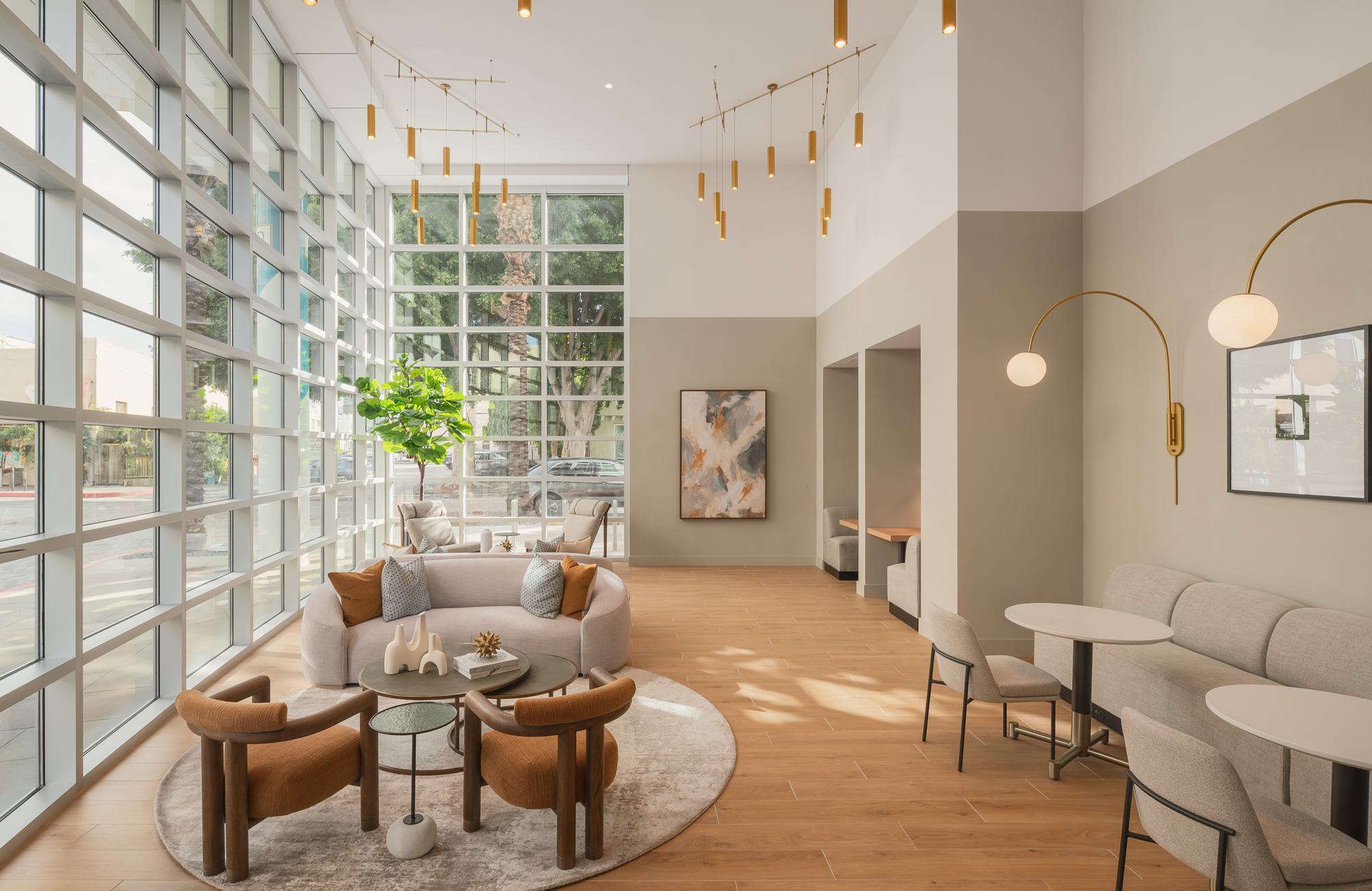 A bright, double-height community lounge with massive grid windows, featuring a modern sofa seating area, cafe tables, and semi-private work booths.