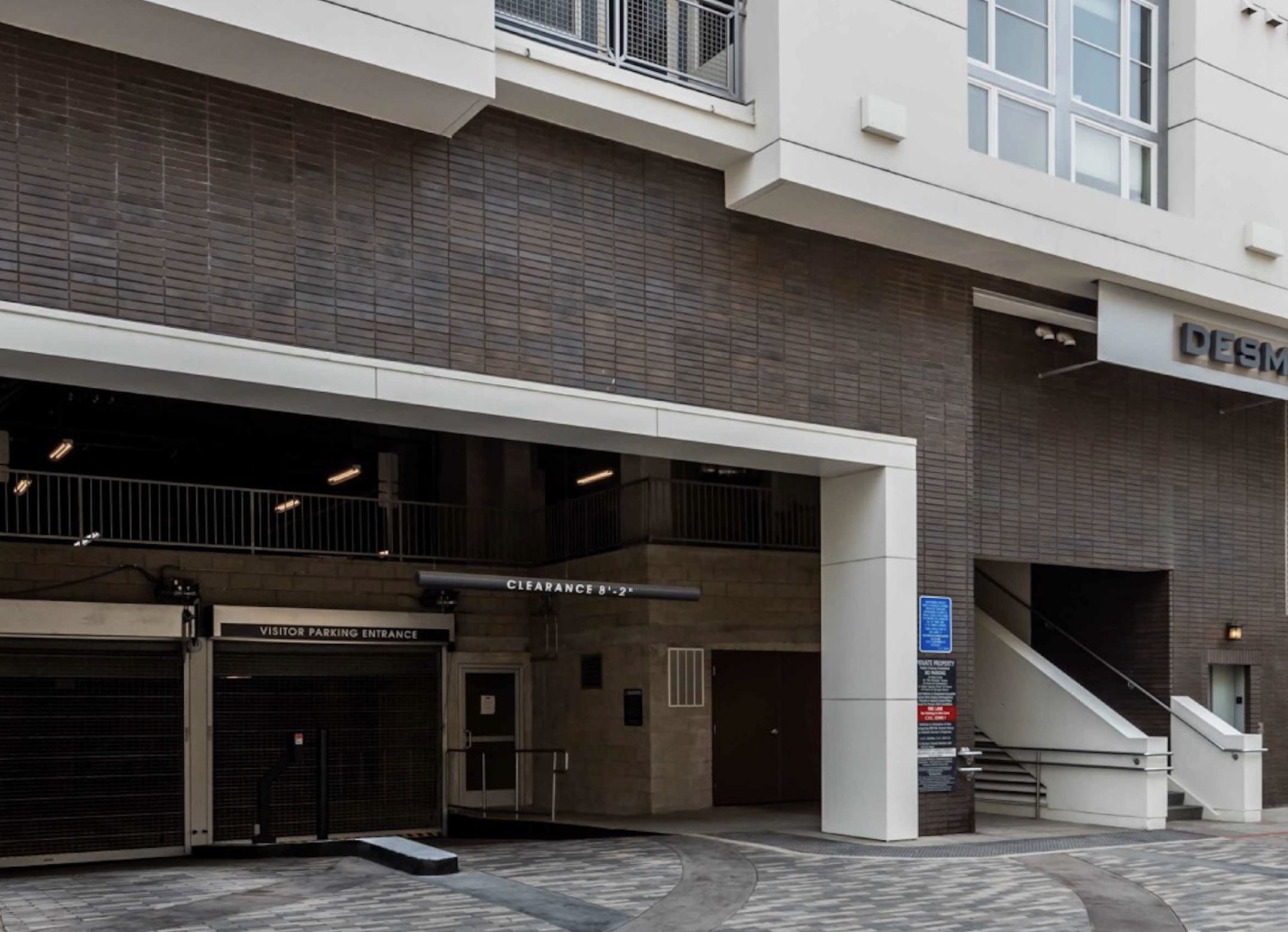 The exterior parking garage entrance at Desmond at Wilshire, featuring a dark brick facade, modern architecture, a clear 8'2" clearance bar, and separate roll-up security gates for the visitor parking entrance