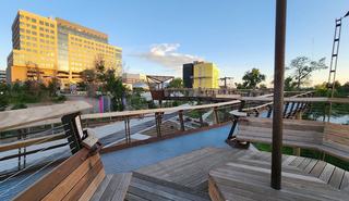 An elevated wooden and metal walkway with built-in benches overlooks the Arkins Promenade in RiNo. Down below, a paved multi-use trail runs parallel to the South Platte River, with modern apartment and office buildings under a clear blue sky in the background.