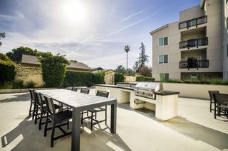 A large modern outdoor dining table and chairs next to a built-in grill station in a sunny apartment courtyard.