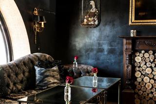  A moody, dark-walled dining corner at Lillie's in the Culver Hotel, featuring a plush patterned sofa, mirrored coffee tables with single red roses in glass vases, a fireplace filled with cut logs, and an eclectic framed painting of a goose wearing a dress.