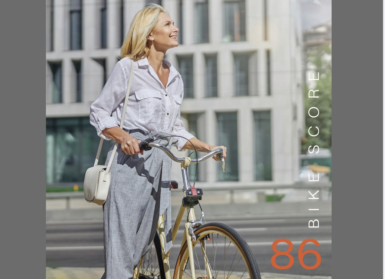 A smiling woman walking next to a bicycle on an urban street with modern buildings in the background. Vertical text reads "BIKE SCORE" next to a large orange number "86."