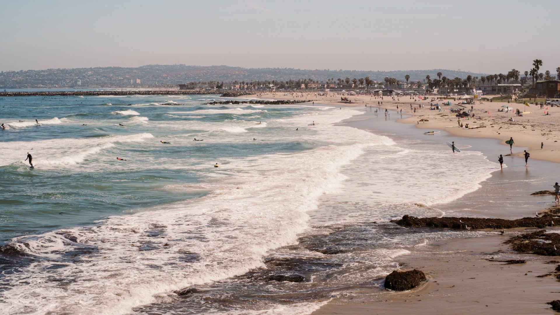 Wide shot of Ocean Beach in San Diego, showing waves crashing on the shore with surfers in the water, people walking along the sandy coastline, and a rocky jetty stretching into the ocean in the background under a clear sky.