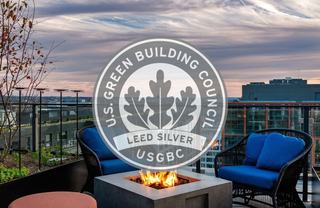 A rooftop terrace at dusk with a burning fire pit and blue lounge chairs, overlaid with the U.S. Green Building Council LEED Silver logo.