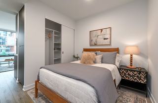 A modern apartment bedroom with a bed, nightstand, and a partially open closet revealing custom built-in vertical shelving and a clothing rod