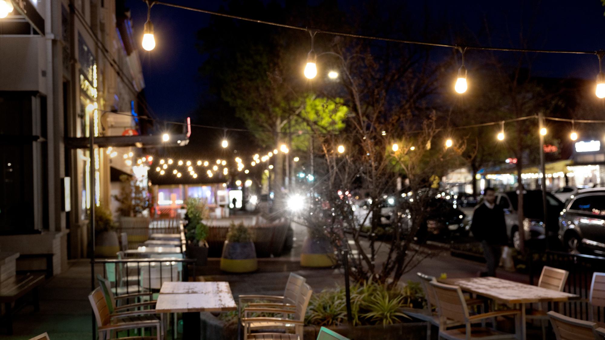 A cozy outdoor restaurant patio at night with wooden tables and chairs, illuminated by overhead strings of warm bistro lights stretching down a city sidewalk.