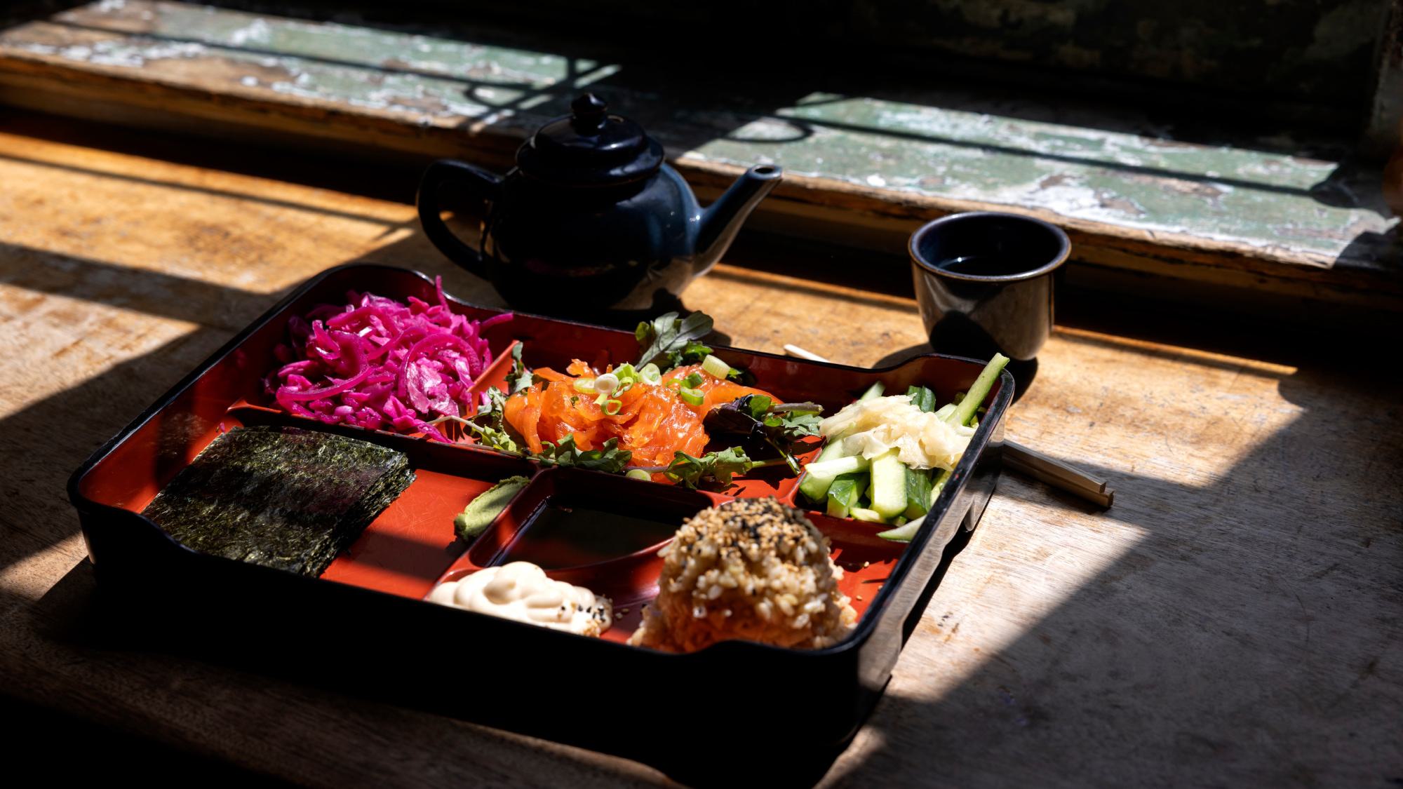apanese bento box filled with salmon, seasoned rice, seaweed sheets, and pickled vegetables sits on a sunlit wooden table next to a dark teapot and cup.