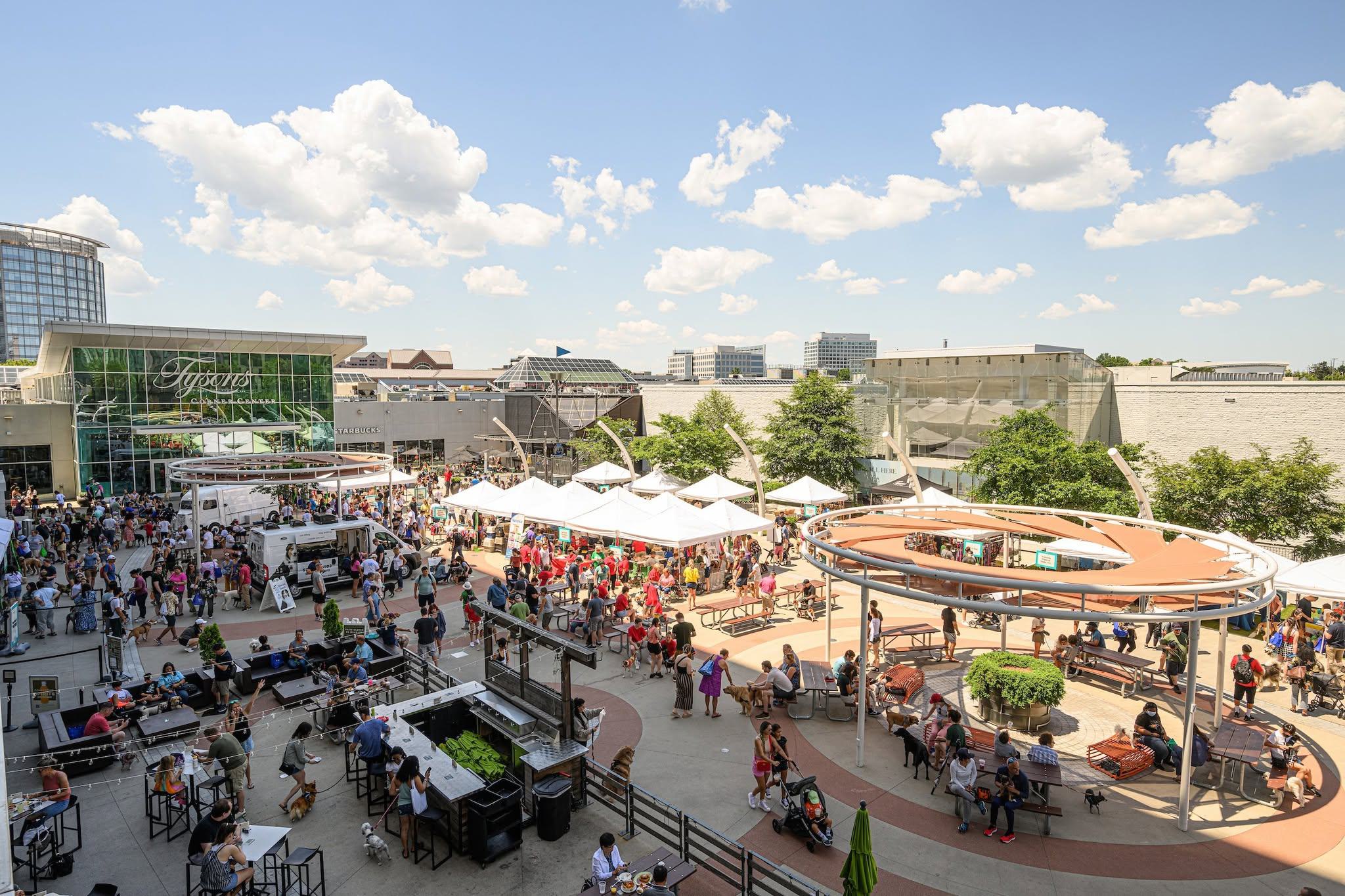 Crowded outdoor pedestrian plaza at Tysons Corner Center featuring food trucks, market tents, and patio seating under a blue sky.