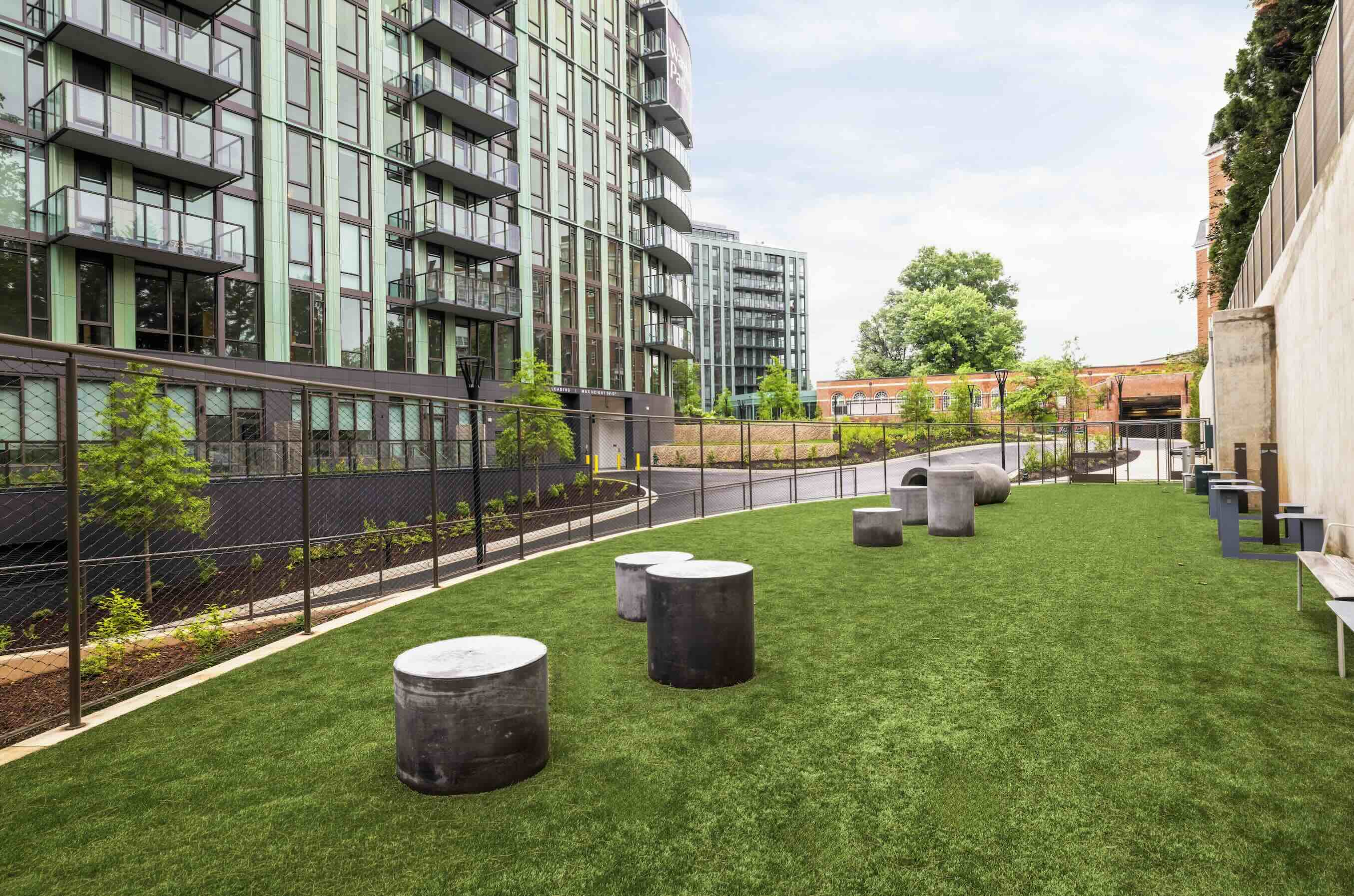 A secure, fenced-in residential dog park with a green turf lawn, concrete agility cylinders, benches, and a dog-friendly water fountain next to a modern tower.