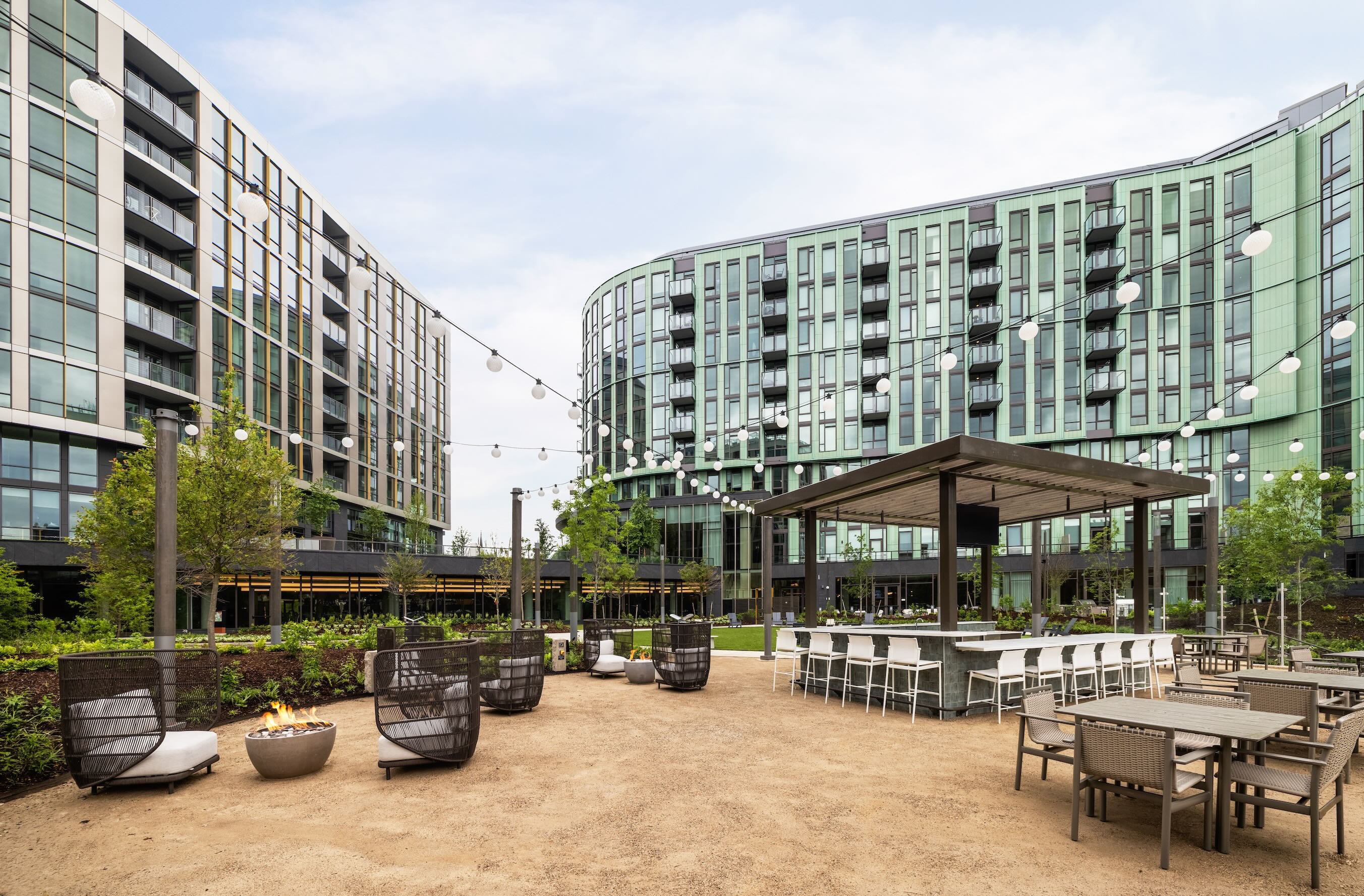 An expansive apartment courtyard at dusk featuring a central bar pavilion under strings of glowing cafe lights, surrounded by modern wicker lounge chairs, fire bowls, and dining tables, with luxury residential buildings in the background.