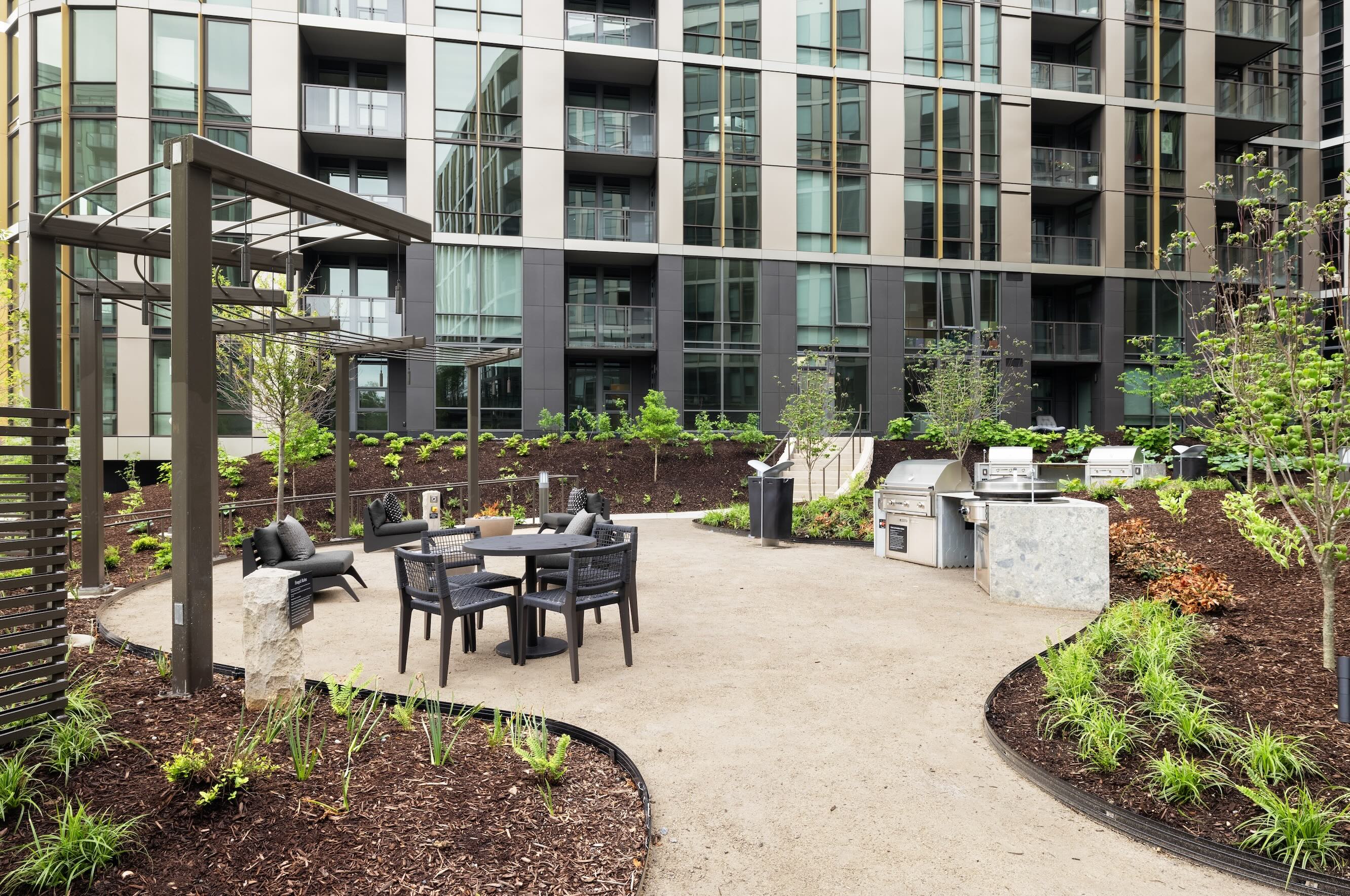 An outdoor courtyard space featuring a stainless-steel BBQ grill station alongside a gravel seating area with a dining table, chairs, and a metal pergola, bordered by mulched garden beds and a modern high-rise.