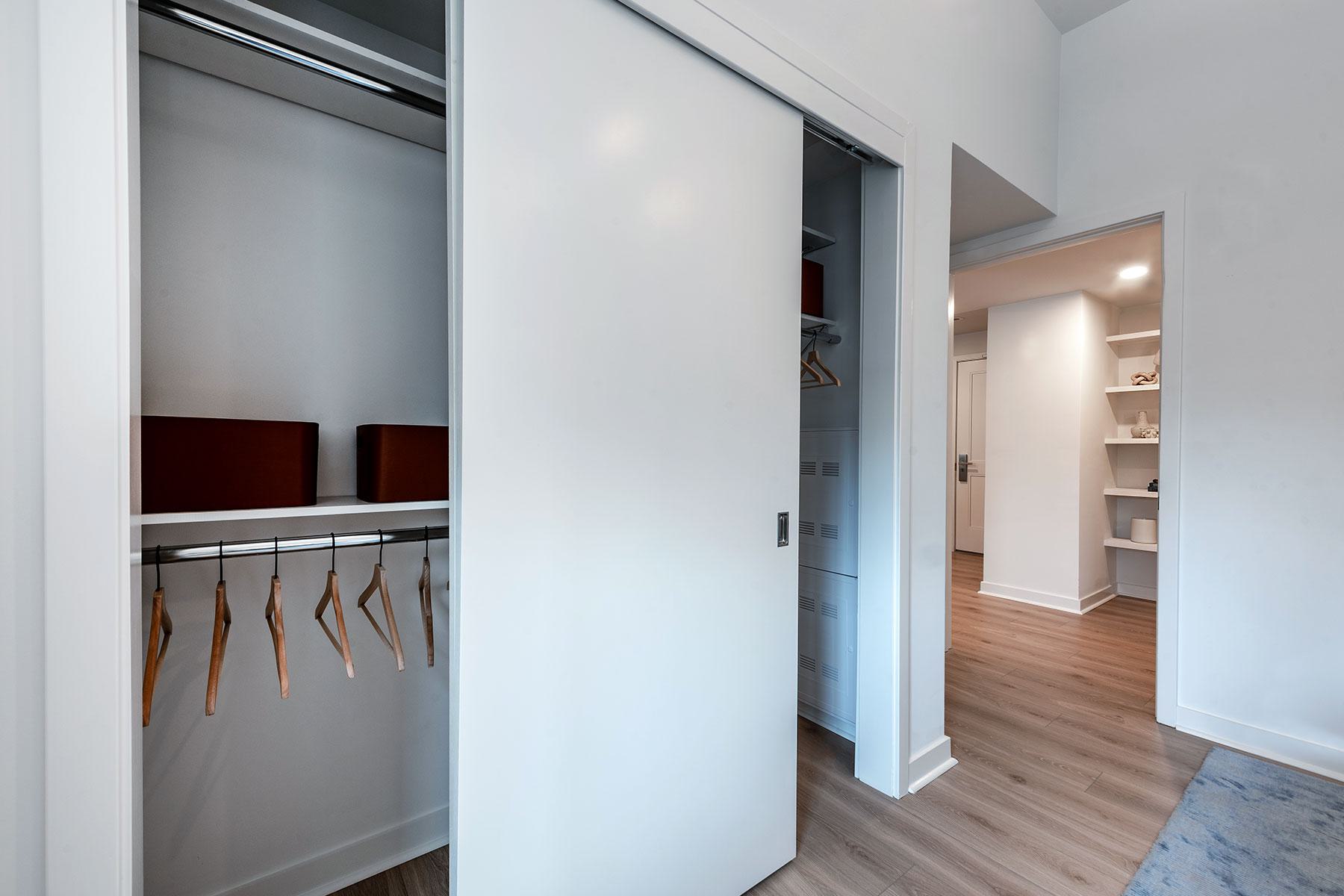 A clean, white closet with a sliding door featuring built-in hanging racks and storage shelves, looking out into an apartment hallway