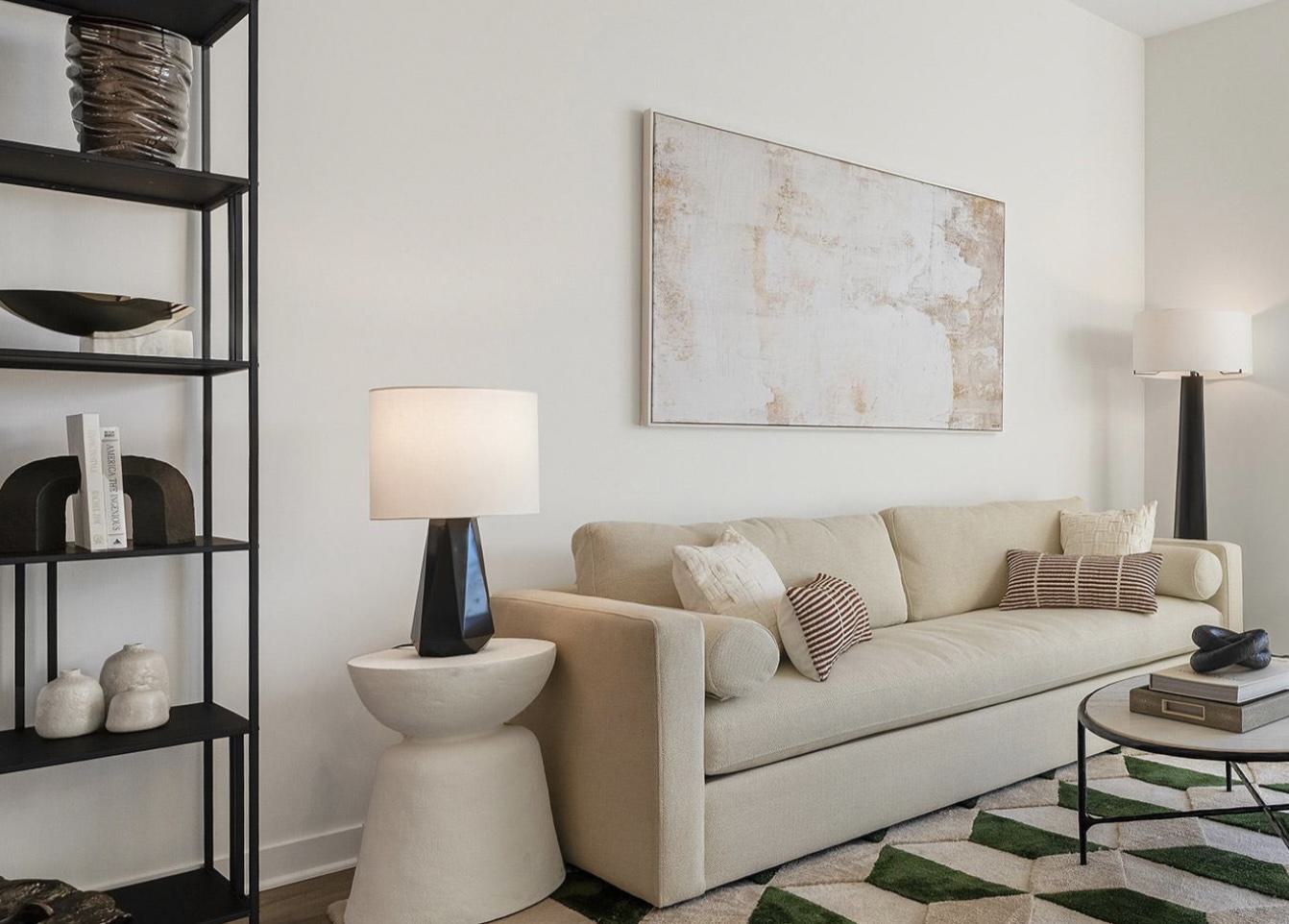 A modern, neutrally decorated den or living room featuring a cream-colored sofa with throw pillows, a geometric patterned rug, a black shelving unit, and a large abstract canvas painting on a white wall.