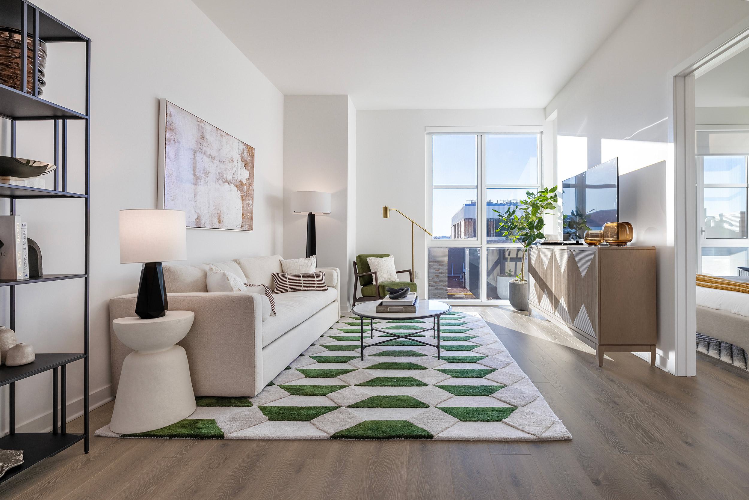 A bright, modern apartment living room with light hardwood floors, a cream-colored sofa, a geometric green and white rug, and large floor-to-ceiling windows letting in bright natural light
