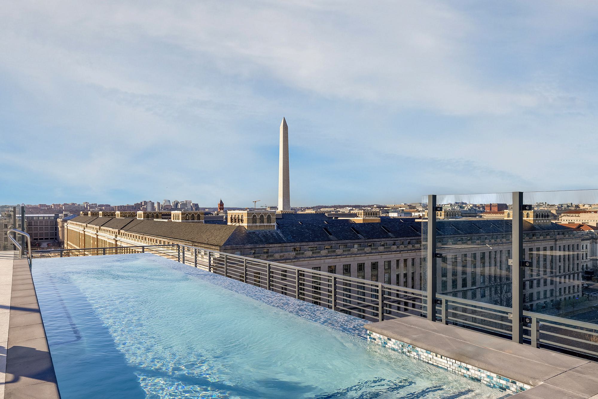 A luxury rooftop swimming pool with a glass safety railing, offering a clear, panoramic view of the Washington Monument and the downtown Washington DC skyline under a partly cloudy sky.