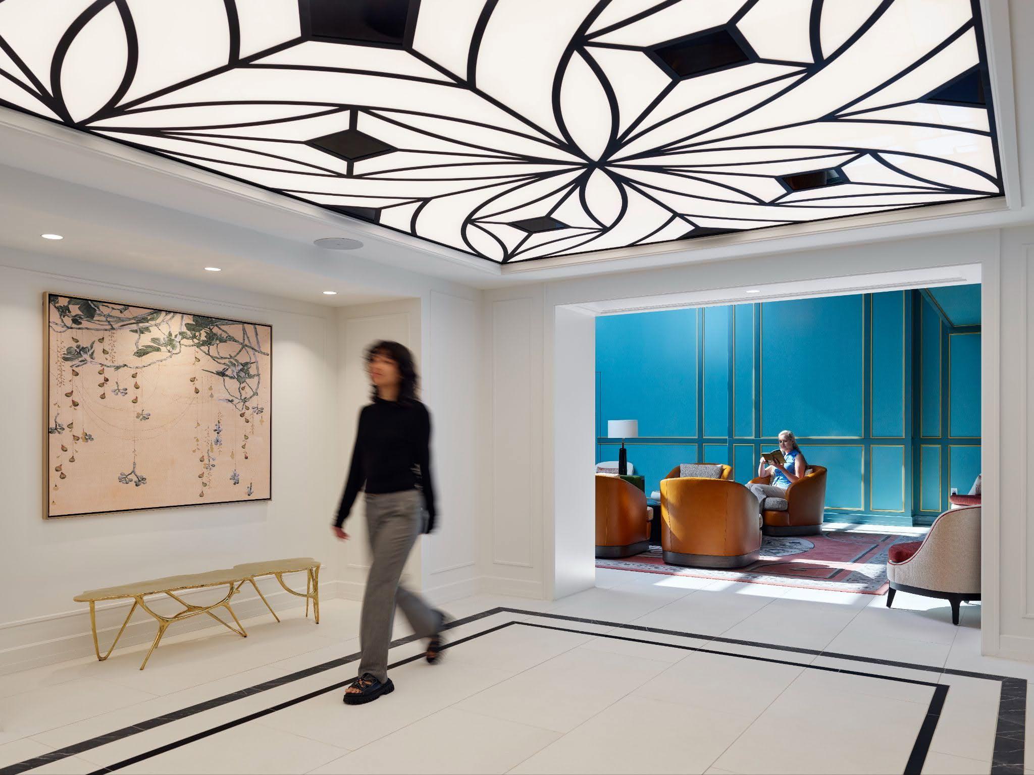 A bright luxury apartment lobby featuring a striking Art Deco-inspired backlit geometric ceiling, white tiled floors, and a view into a lounge with bright teal walls and leather armchairs.