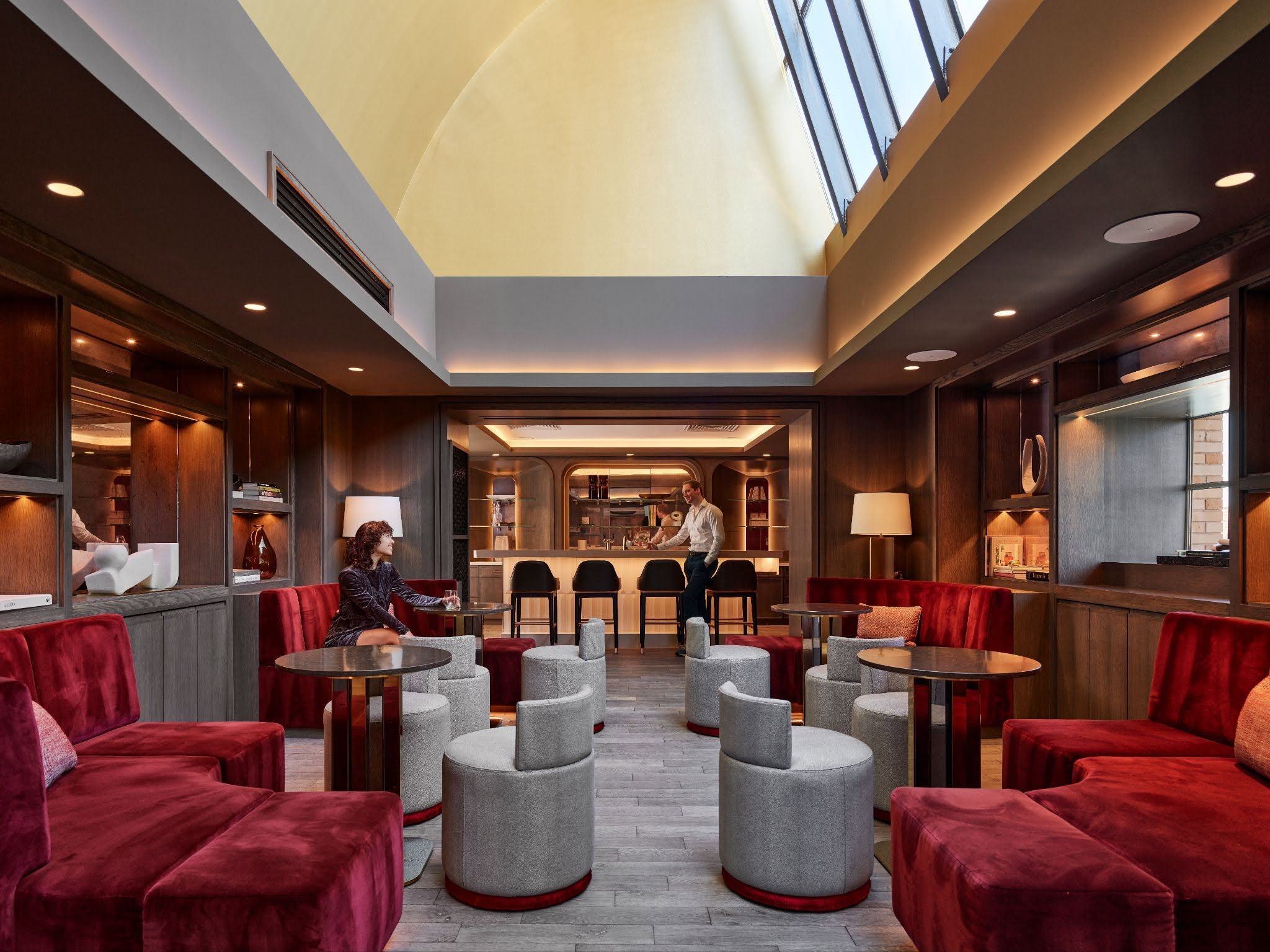 A moody speakeasy bar and lounge with red velvet seating, dark wood paneling, and an upper vaulted atrium ceiling with large skylights throwing warm light over the space.